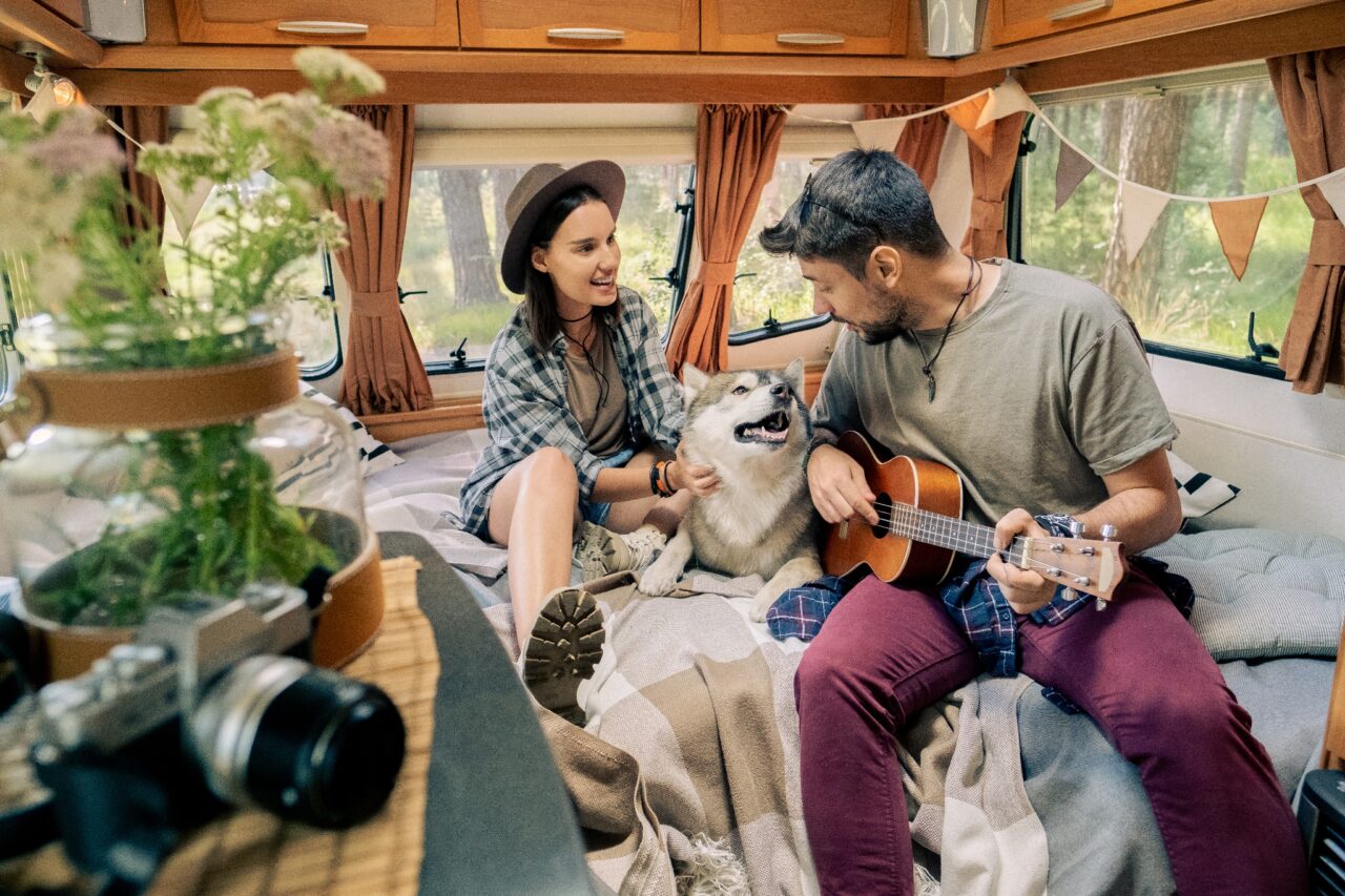 Paar sitzt in schön dekoriertem Campingfahrzeug mit ihrem Husky auf dem Bett, Mann spielt Gitarre.
