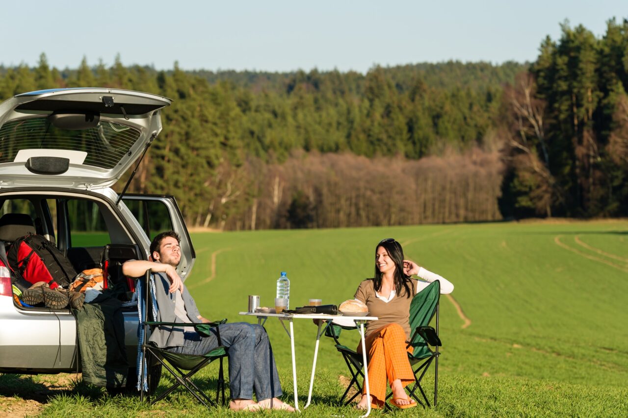 Ein Paar sitzt auf Campingstühlen in der Natur vor einem Auto mit geöffnetem Kofferraum.