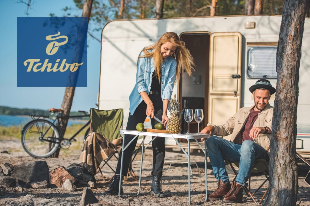 Junges Paar ist vor einem Campingfahrzeug zu sehen, der Mann sitzt auf einem Campingstuhl, die Frau schneidet stehend Avocados auf einem Campingtisch. Oben links ist das Logo von Tchibo zu sehen.